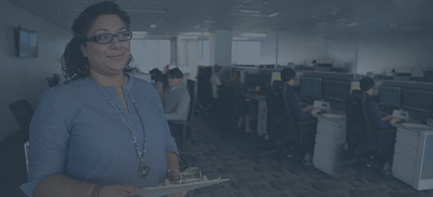 A woman is being interviewed while standing at the front of an office filled with cubicles.