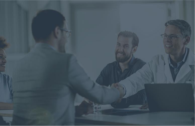 A doctor at a table with a computer and two colleagues is shaking hands with a business consultant.
