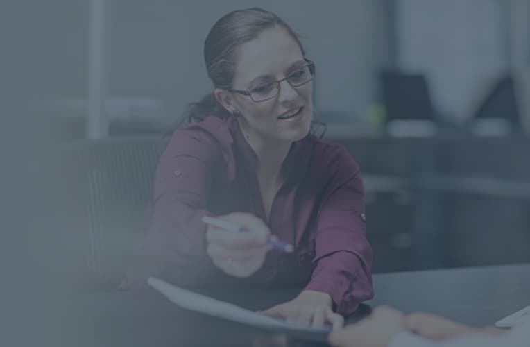 A business woman in glasses is holding out a document and pen for someone to sign.