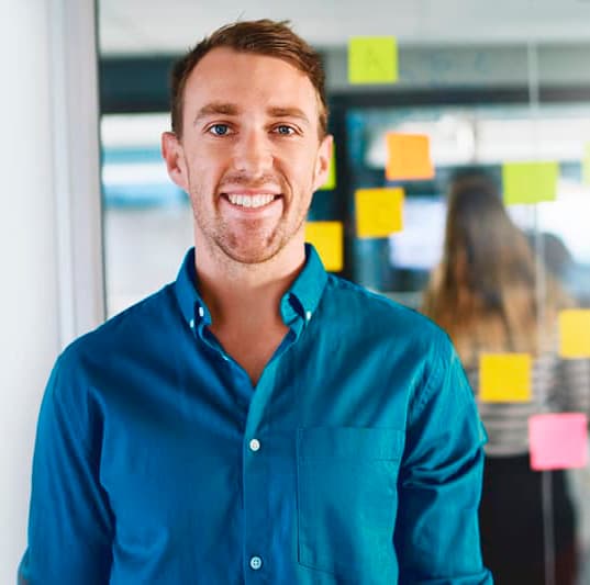 A front-facing, upper body shot of a man standing in front of an office window with post-it notes.